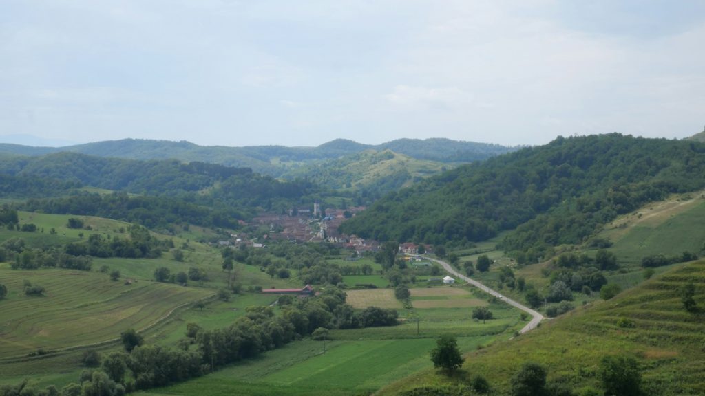 Sat transilvănean într-o vale verde, cu dealuri, câmpuri și un drum care intră în sat